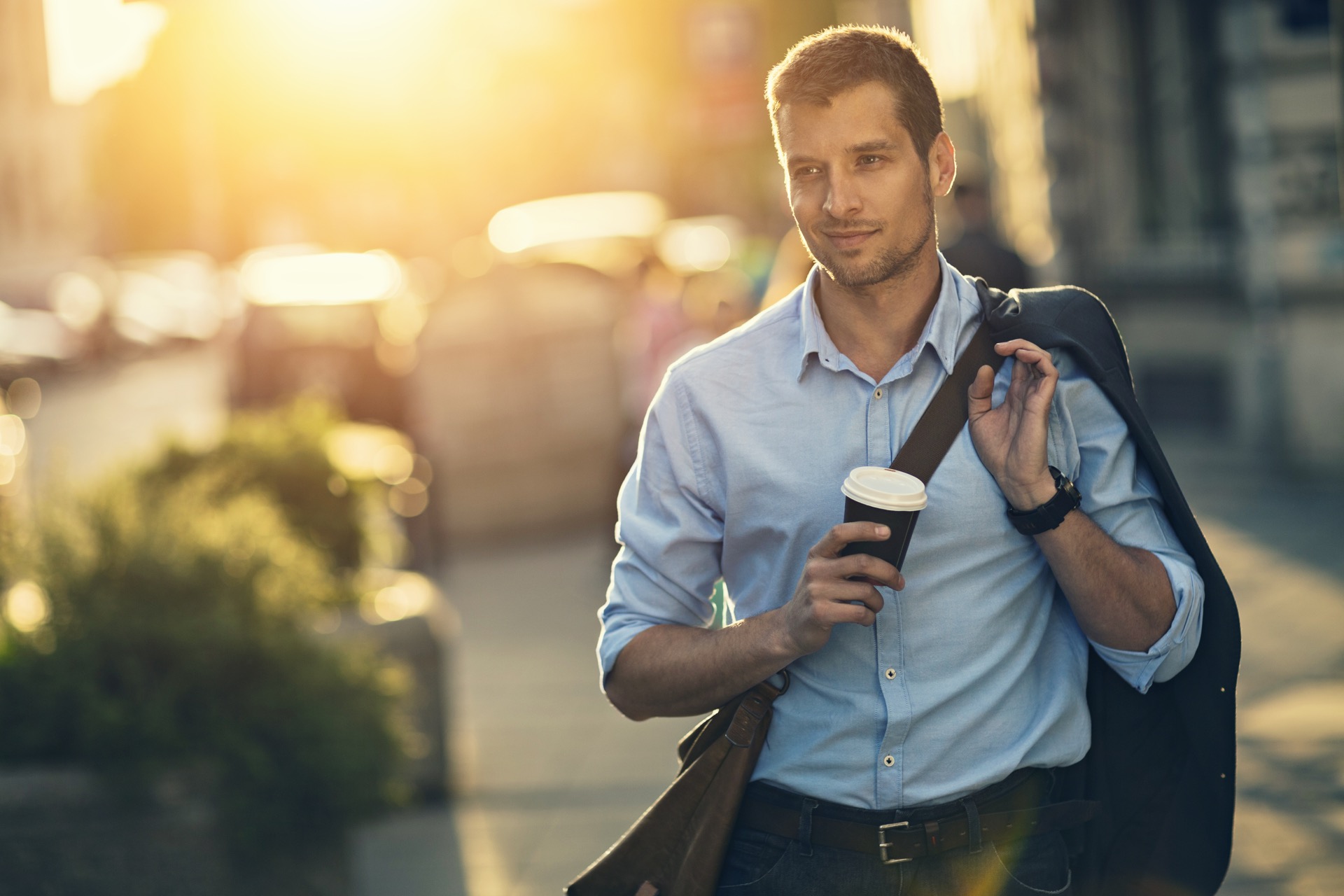 Picture of a young handsome business man having take away coffee walking home from work in the city. He is dressed in a blue shirt with the sleeved rolled up, carrying his jacket over his shoulder. He also has a brown leather bag over his other shoulder and wearing dark jeans with a brown belt. In the background are bushes, other people walking and city buildings with the sun setting over the roof tops.