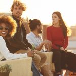 Portrait of smiling young woman wearing sunglasses while sitting by man during rooftop party. Multi-ethnic male and female friends spending leisure time. They are wearing casuals on sunny day.